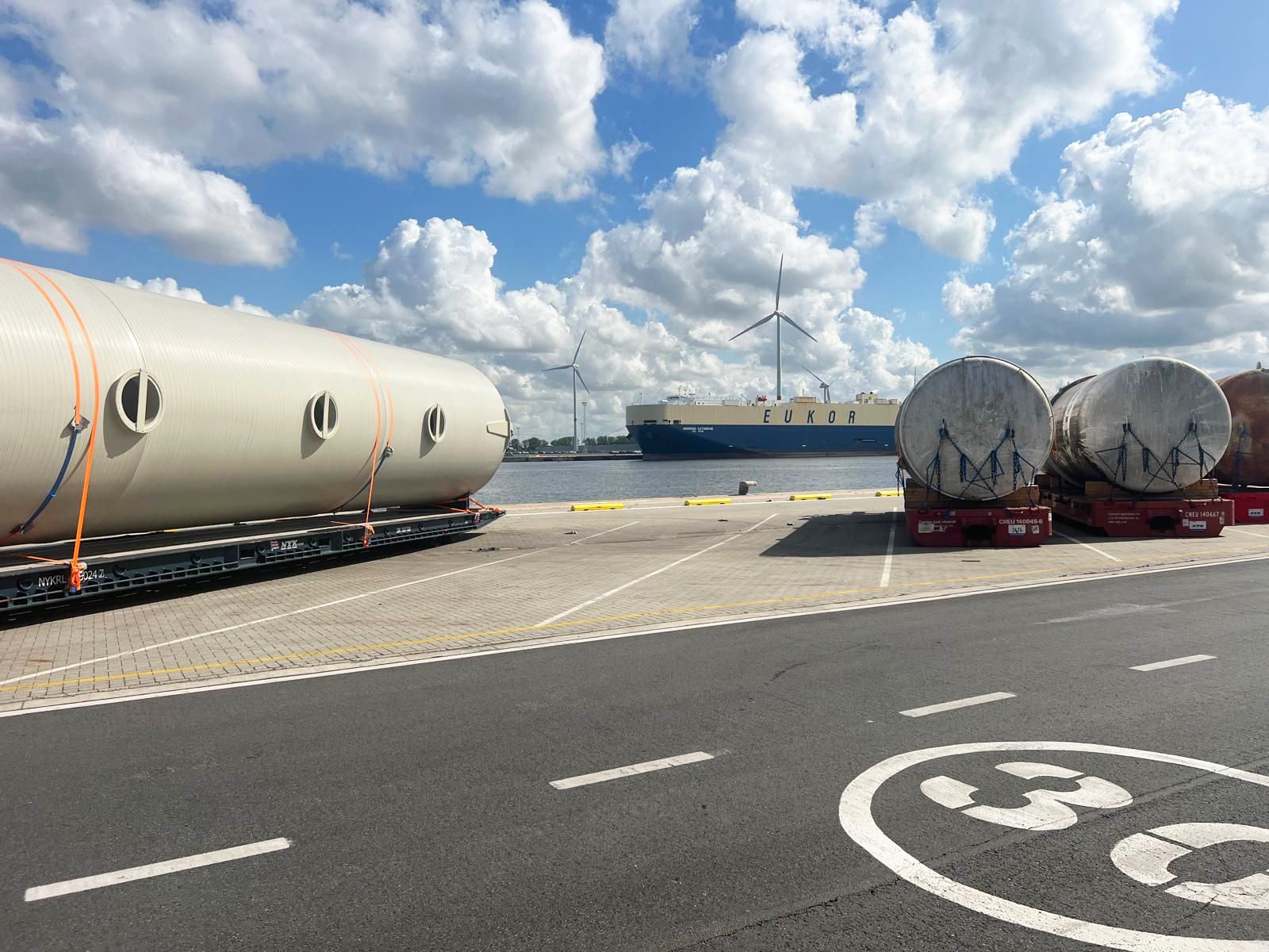 Cylindrical industrial tanks secured on flatbed trailers at Zeebrugge port with cargo ship and wind turbines in background, 2025